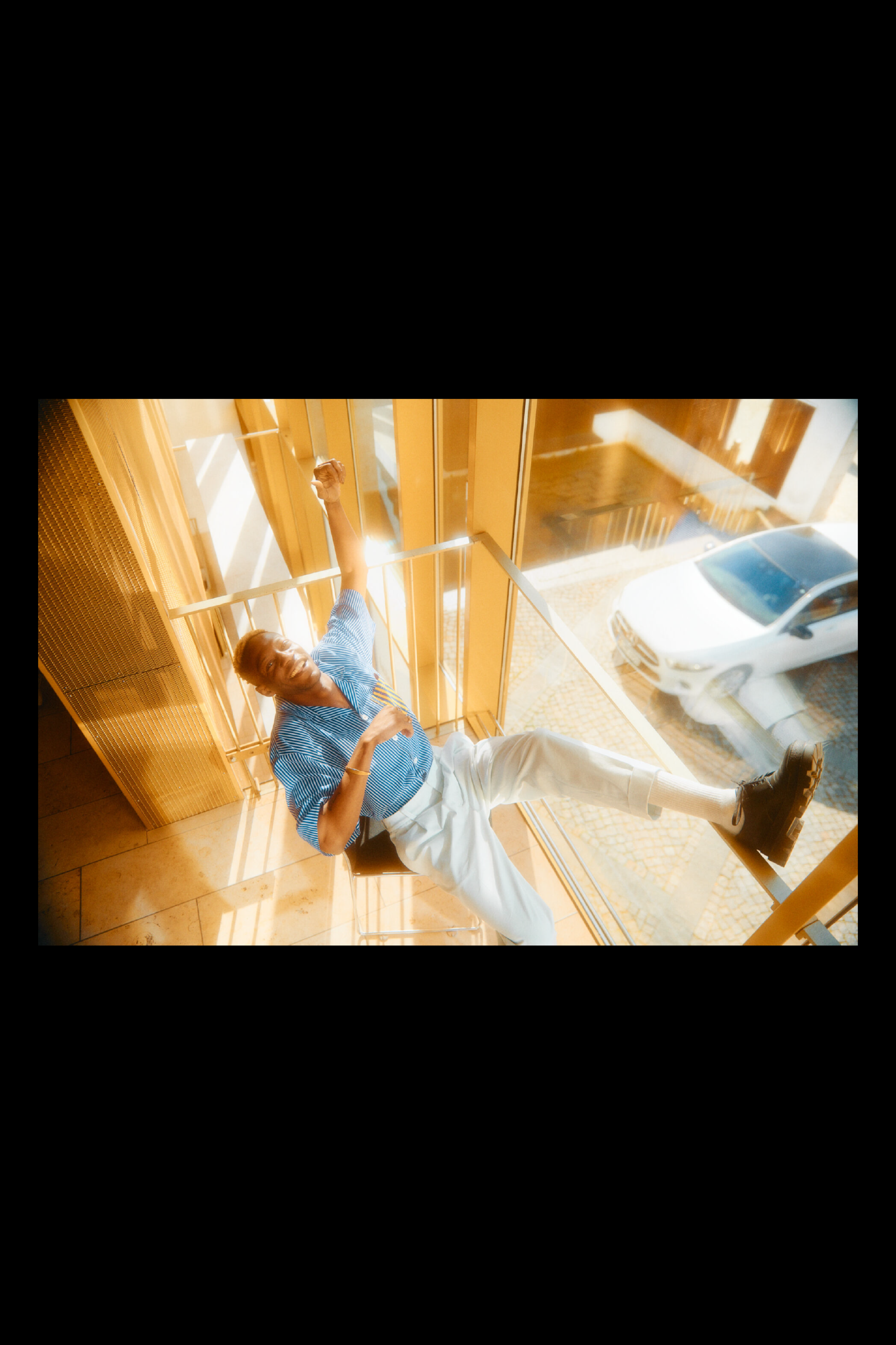 Photography of a man sitting on a chair at a huge window, outside is a white car